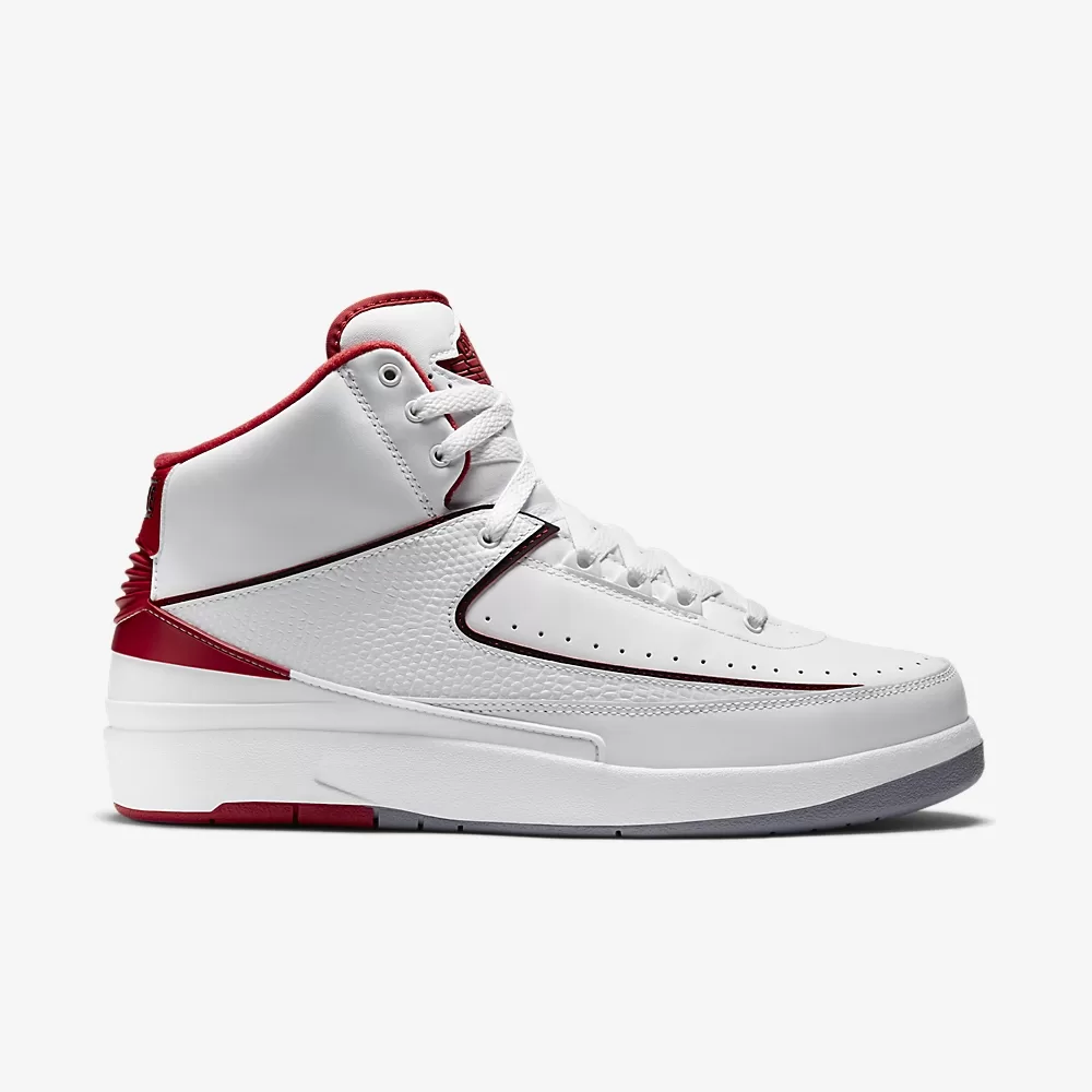 A close-up photo of the Air Jordan 2 Retro "White Fire Red Black Cement" sneaker. The sneaker has a white leather upper with black and fire red accents. The Nike Air logo is embroidered on the tongue and heel in black. The midsole is white and the outsole is black and fire red.