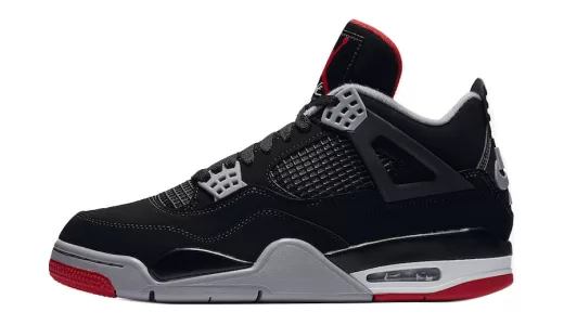 A close-up photo of the Air Jordan 4 "Bred" Reimagined sneaker. The sneaker has a black leather upper with red accents on the tongue, Wings logo, and eyelets. The Nike Air logo is embroidered on the heel in black. The midsole is white and the outsole is black and red.